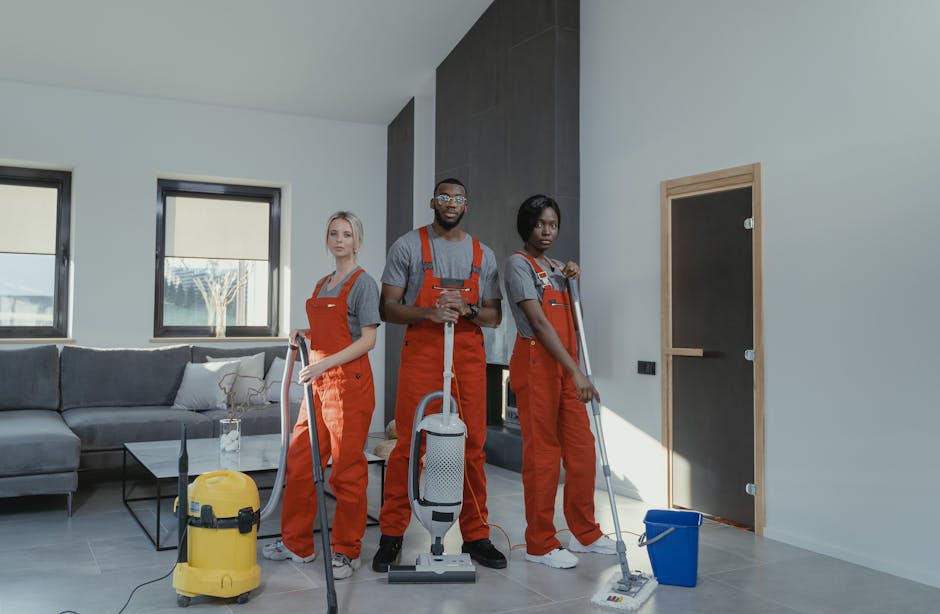 A person wearing beige pants and bright blue athletic shoes is sweeping a polished wooden floor with a broom, removing dust and debris. The scene is well-lit, highlighting the smooth, shiny surface of