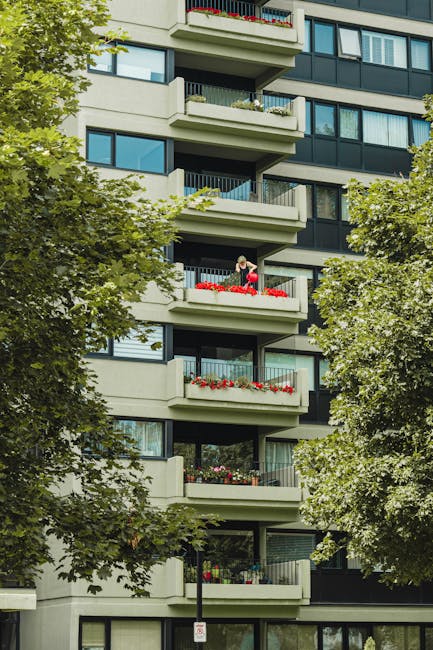 A multi-story residential building with several balconies, each featuring glass sliding doors and concrete railings. Some balconies are decorated with red and pink flowers in planters, and a person is visible tending to plants on one of them. The building's facade is light-colored with black window frames, and lush green trees partially obscure the lower floors, framing the scene. The overall atmosphere is bright and clean, highlighting the building's modern design and maintained appearance, consistent with high-quality domestic cleaning standards promoted by Earls Court Cleaner for deep cleaning and surface sanitisation of residential exteriors.